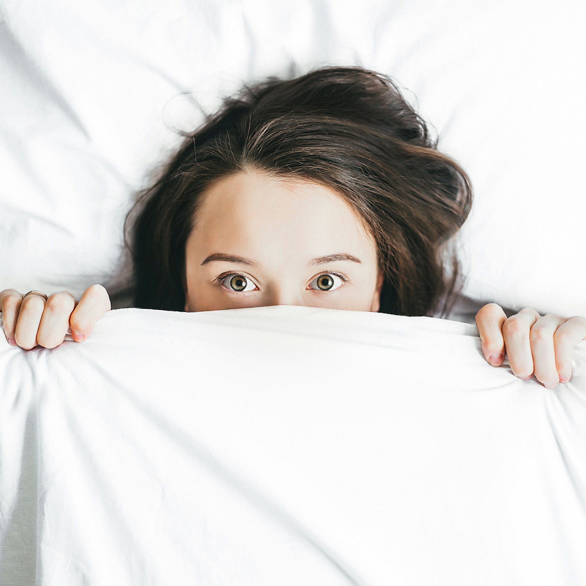 A woman lying in white bedding, awake, watching with wide eyes over the duvet.