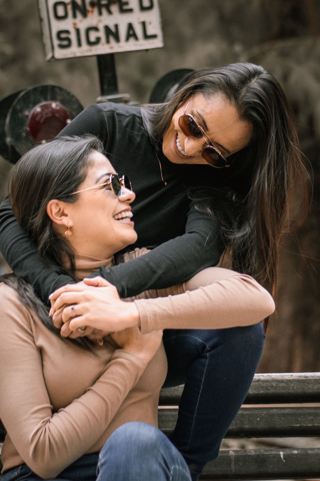 Two women laughing together on a bench, one with an arm wrapped around the other.