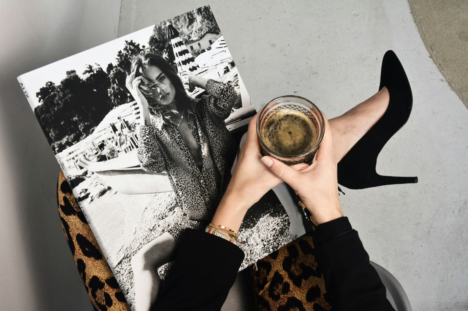 An editorial flatlay: a black-and-white fashion magazine, an espresso held in both hands, a heel resting alongside.