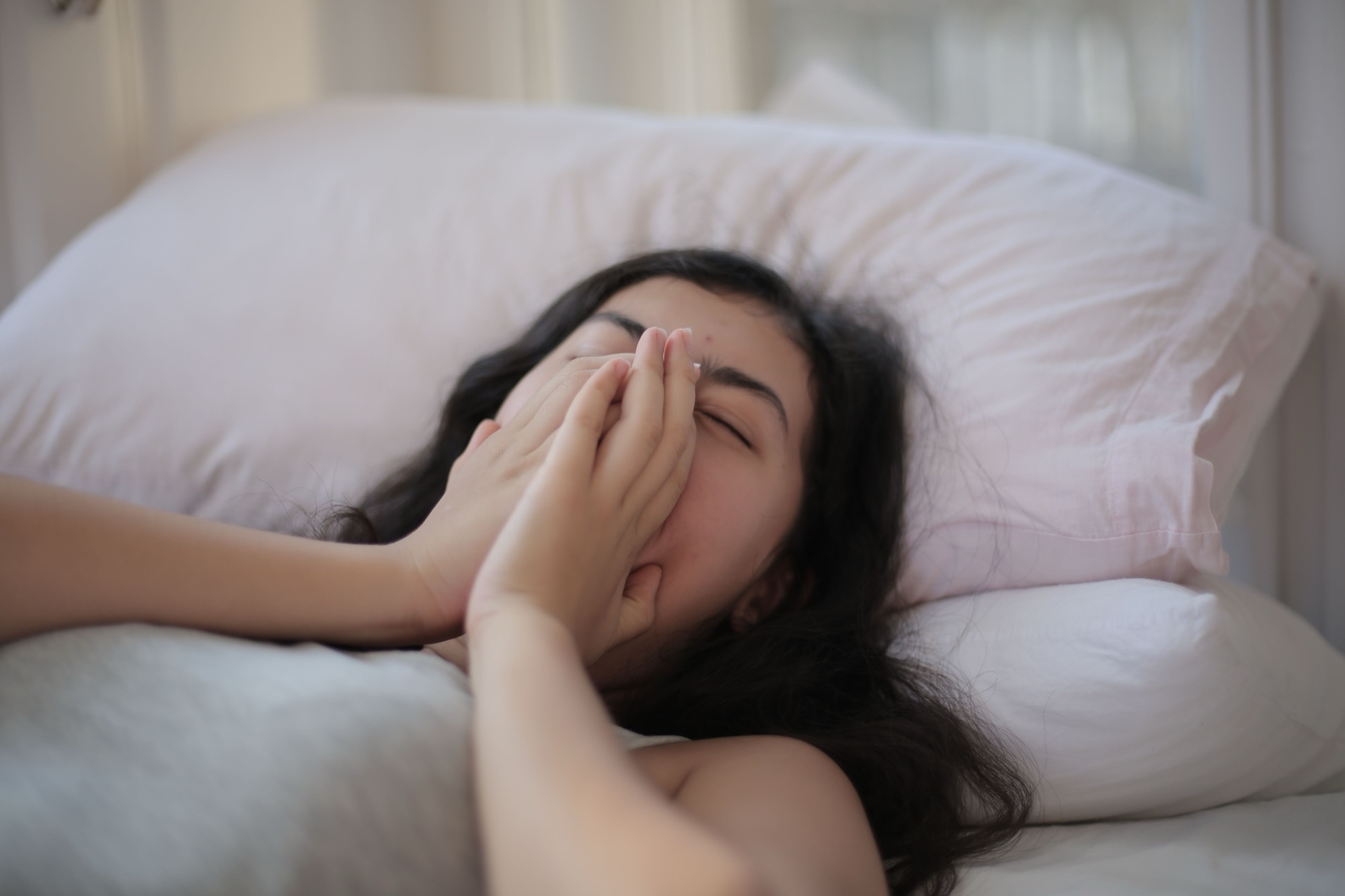 A woman lying in bed, hands over her face, awake and tired in soft morning light.
