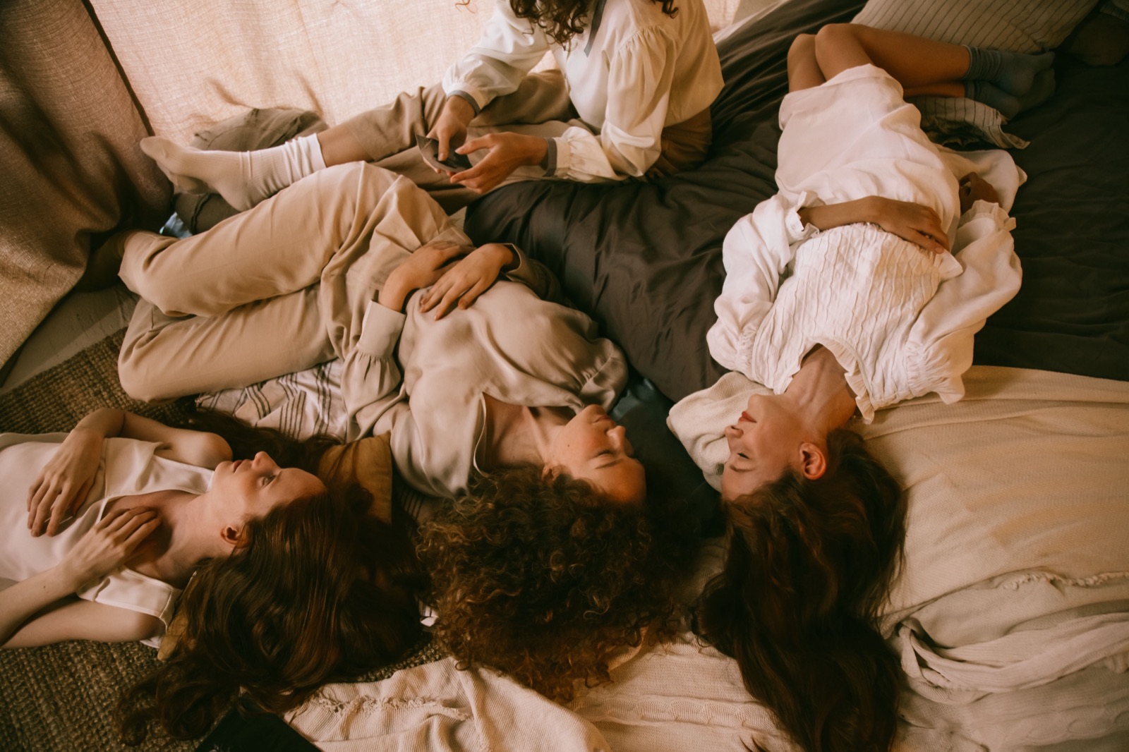 Four women in soft neutral loungewear lying together in a circle on warm bedding, at rest.