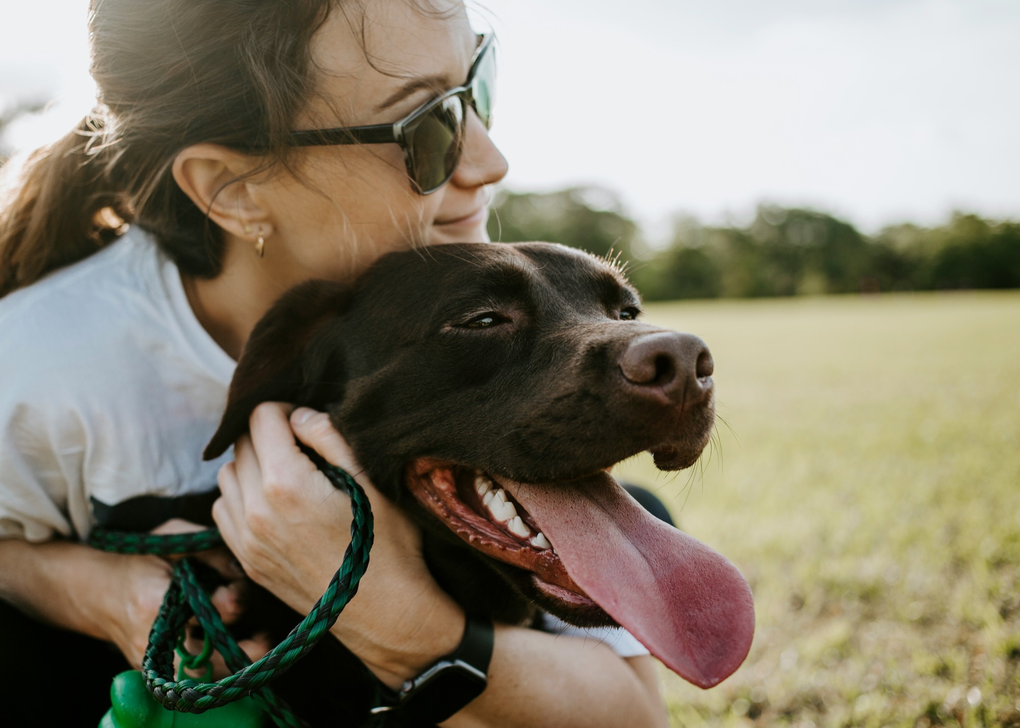 A smiling woman outdoors in warm evening light, her arms around a happy dog.