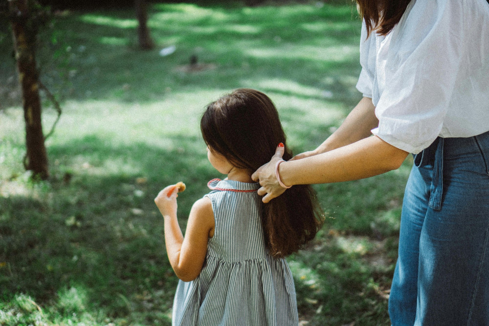 A mother's hand tucking her small daughter's hair behind her ear in a sunlit garden, both seen from behind.