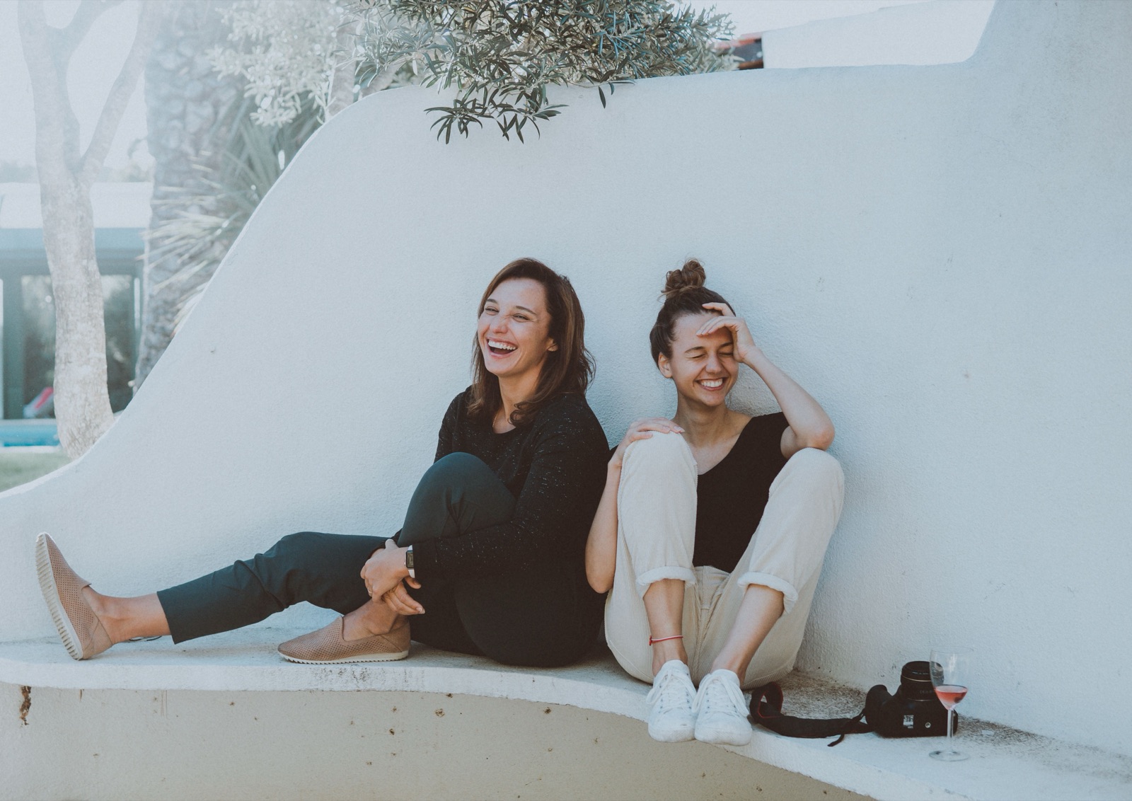 Two women sitting side by side against a white wall, laughing in bright daylight.