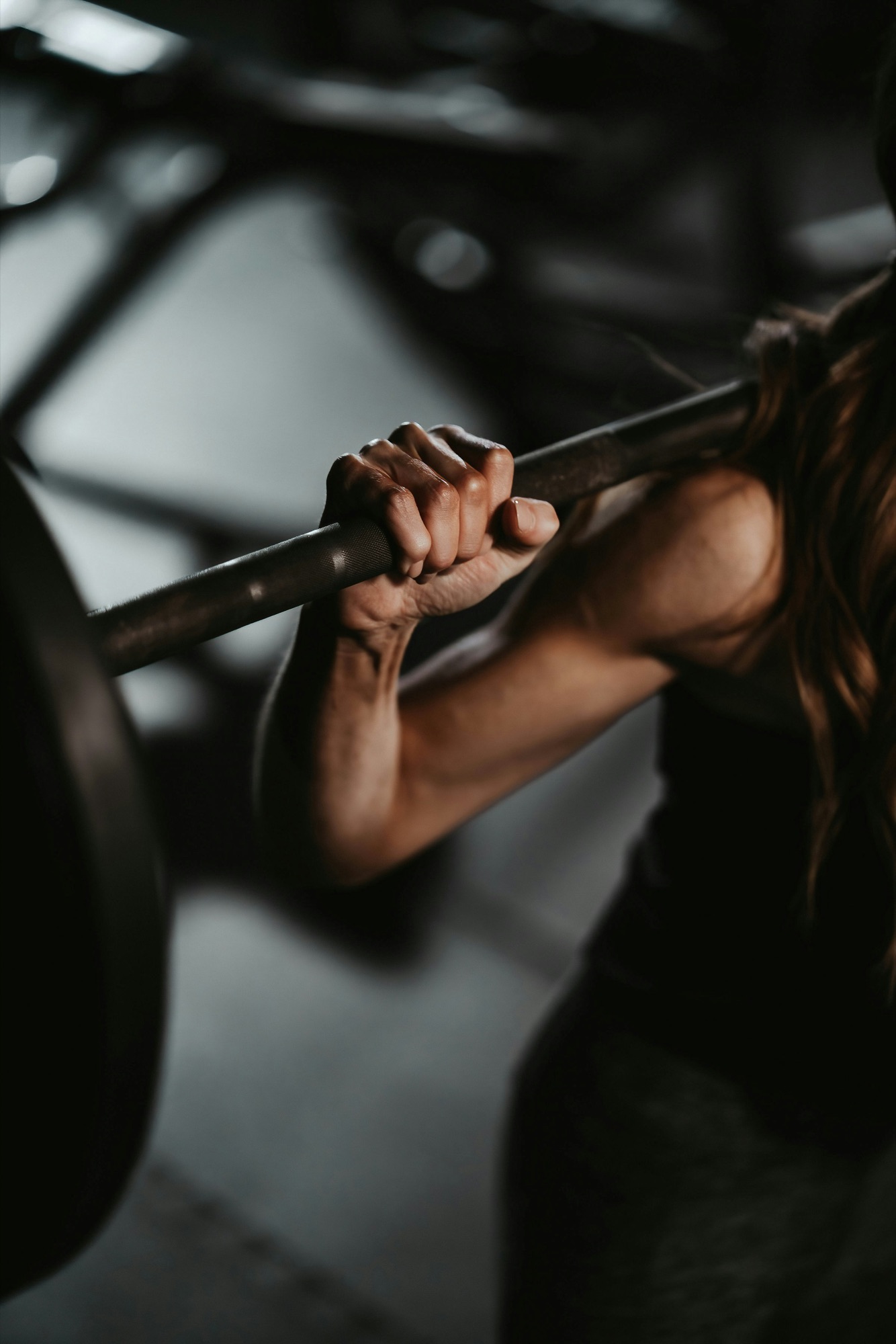 A woman's hands gripping a loaded barbell across her shoulders in a dim gym, her face turned away.