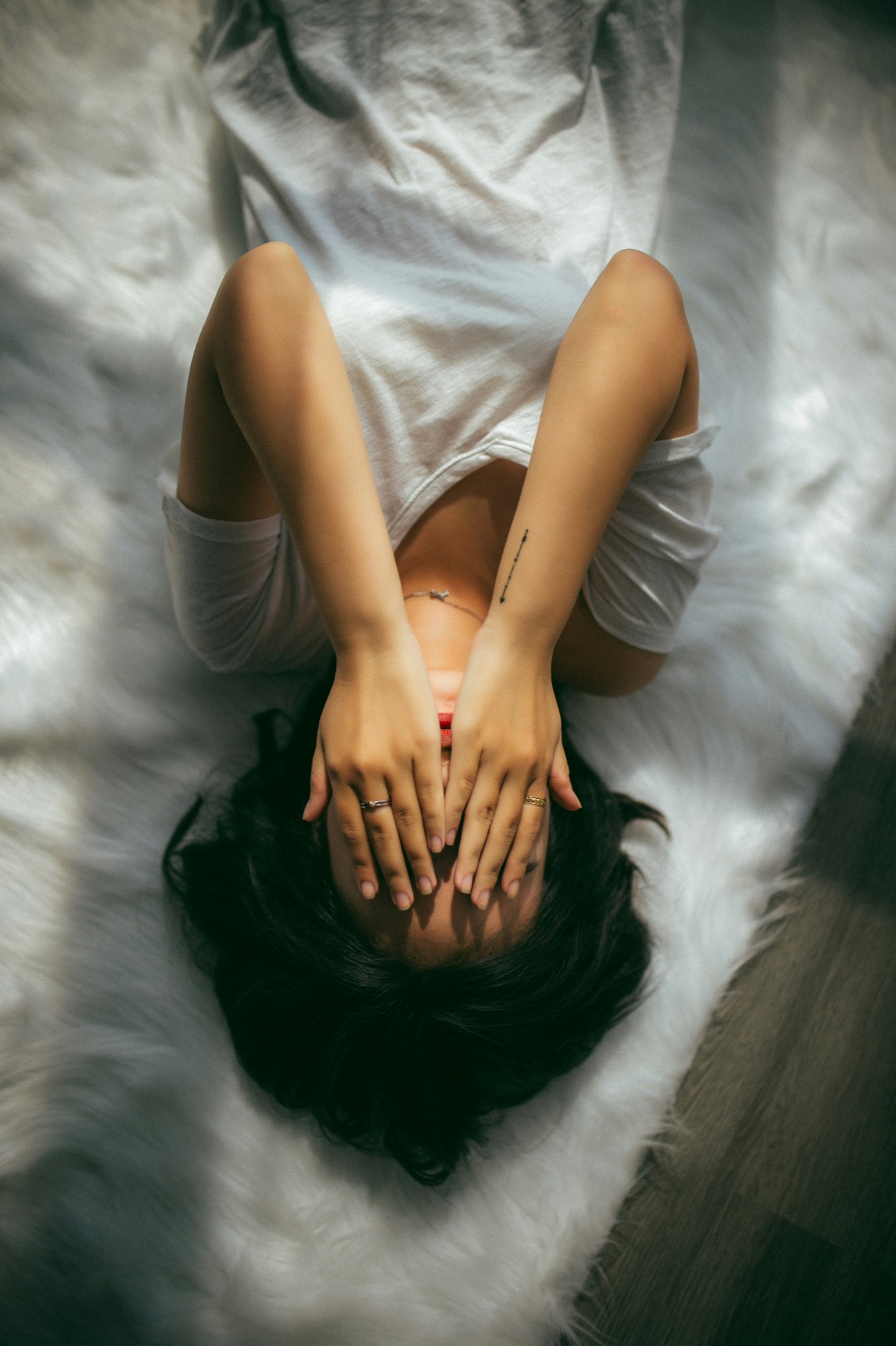 A woman lying on white bedding with both hands covering her face.