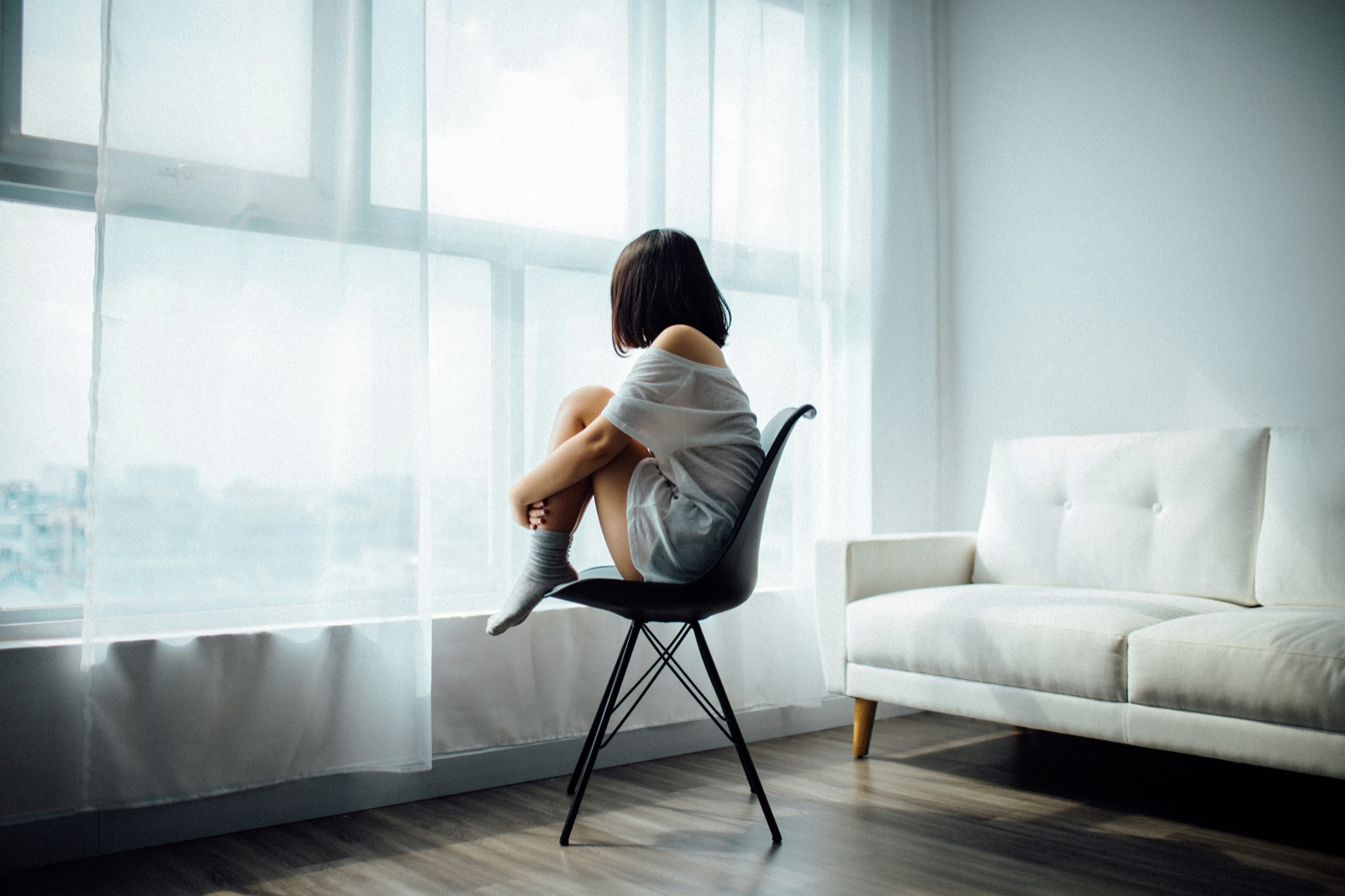 A woman sitting curled on a chair by a tall bright window, looking out over the city in a calm, airy room.