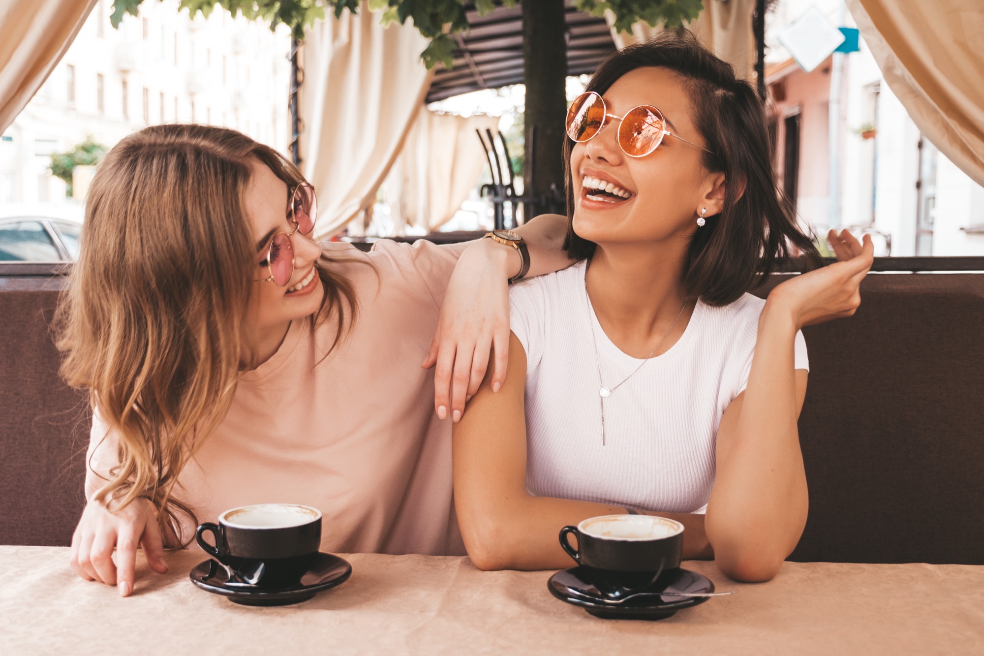 Two women laughing together over coffee at a cafe table, one with an arm around the other's shoulder.