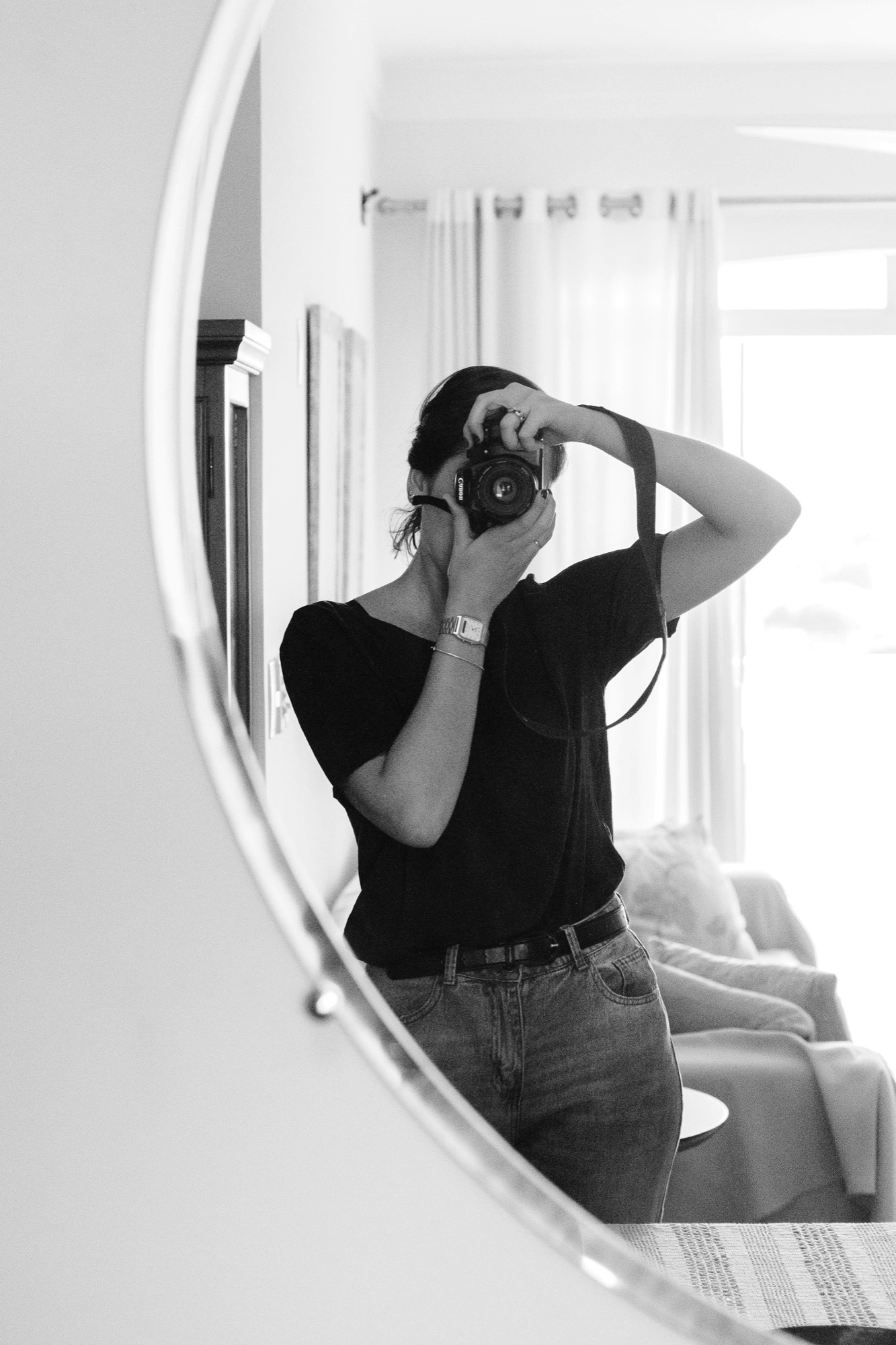 A woman photographing her own reflection in a round mirror, in black and white.