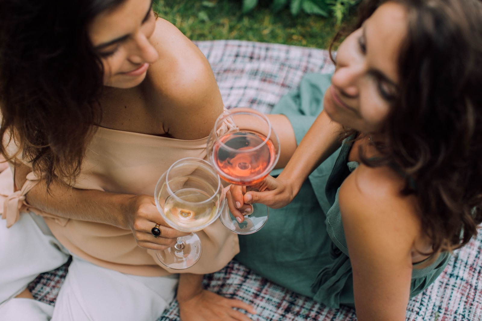 Two women toasting on a picnic blanket in a garden, one glass of rose and one of white wine.